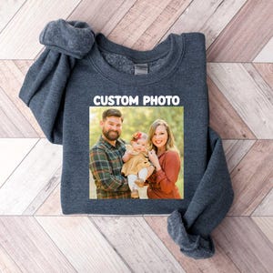 Puede incluir: Una sudadera gris oscuro con una foto familiar rectangular. La imagen muestra a un hombre, una mujer y un beb&eacute;. El texto "CUSTOM PHOTO" est&aacute; encima de la imagen en blanco. La sudadera est&aacute; sobre una superficie de madera.