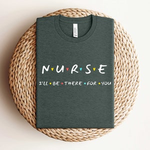Puede incluir: Camiseta gris oscuro con el texto "NURSE" en letras de colores con corazones entre cada letra. El texto "I'll be there for you" est&aacute; impreso en negro debajo.
