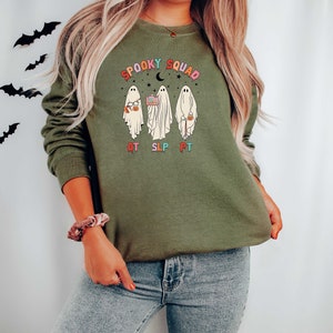 May include: A green sweatshirt with a graphic of three ghosts wearing costumes and holding Halloween treats. The text "Spooky Squad" is above the ghosts and the text "OT SLP PT" is below the ghosts.
