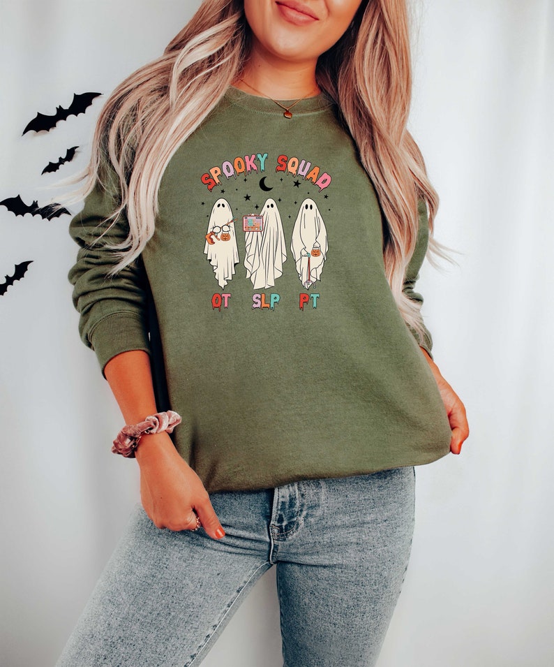 May include: A green sweatshirt with a graphic of three ghosts wearing costumes and holding Halloween treats. The text "Spooky Squad" is above the ghosts and the text "OT SLP PT" is below the ghosts.