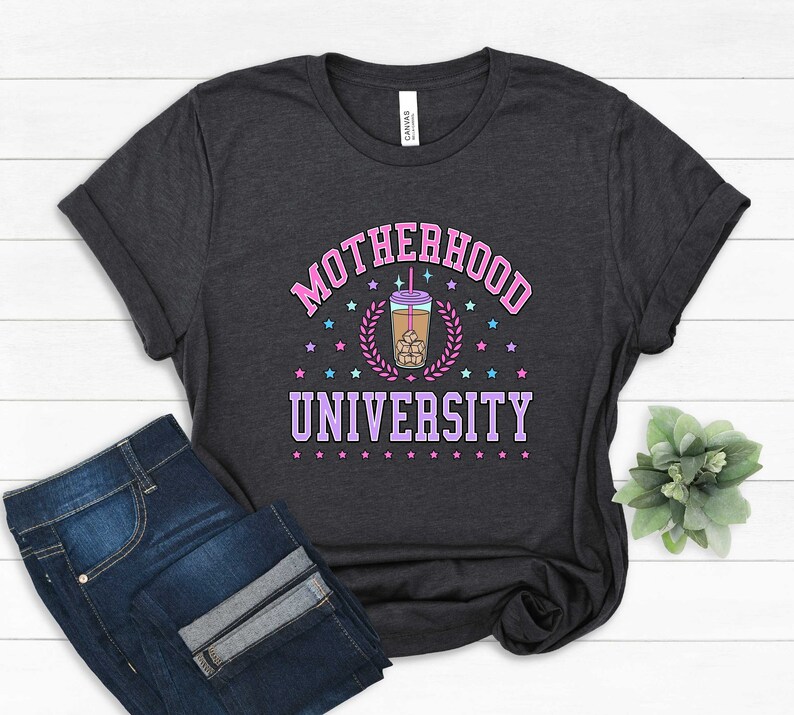 May include: A dark gray t-shirt with the text "MOTHERHOOD UNIVERSITY" in pink and purple lettering. A cartoon illustration of a cup of iced coffee with a straw and ice cubes is in the center of the text. The text is surrounded by small pink and blue stars.