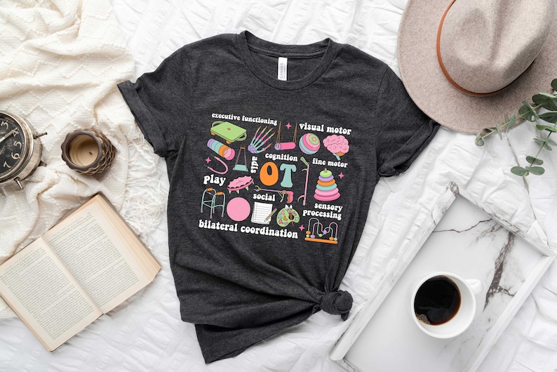 May include: A dark gray t-shirt with a colorful graphic design featuring the word "OT" and illustrations of various toys and activities. The text around the graphic includes "play", "social", "cognition", "sensory processing", "bilateral coordination", "executive functioning", "visual motor", and "fine motor".