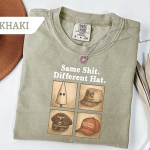 Peut inclure: T-shirt kaki avec le texte "Same Shit. Different Hat." et quatre illustrations de chapeaux diff&eacute;rents. Un collier argent&eacute; avec un pendentif repose sur le t-shirt. Le t-shirt est &eacute;tiquet&eacute; "Comfort Colors."