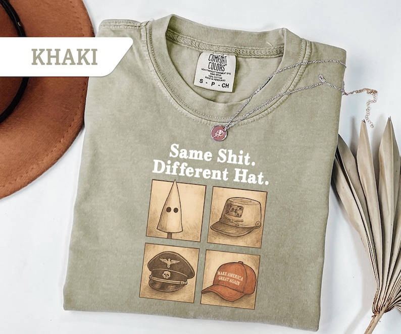 Peut inclure: T-shirt kaki avec le texte "Same Shit. Different Hat." et quatre illustrations de chapeaux diff&eacute;rents. Un collier argent&eacute; avec un pendentif repose sur le t-shirt. Le t-shirt est &eacute;tiquet&eacute; "Comfort Colors."