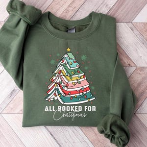 May include: Green sweatshirt with a Christmas tree design made of books and string lights. The text "ALL BOOKED FOR Christmas" is printed below the tree. The sweatshirt is folded on a wooden surface.