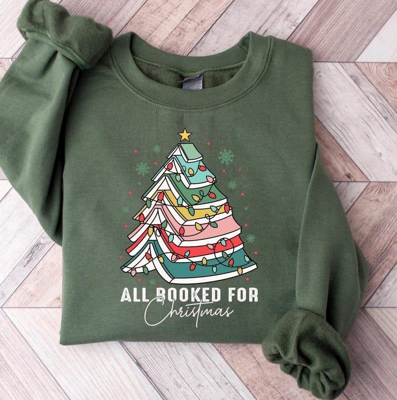 May include: Green sweatshirt with a Christmas tree design made of books and string lights. The text "ALL BOOKED FOR Christmas" is printed below the tree. The sweatshirt is folded on a wooden surface.