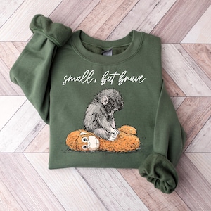 May include: A green sweatshirt with the text "small, but brave" in white script. The graphic features a detailed illustration of a monkey hugging a stuffed animal. The sweatshirt is folded on a wooden surface.