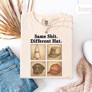Peut inclure: T-shirt de couleur ivoire avec l'inscription "Same Shit. Different Hat." et quatre illustrations de chapeaux. Le t-shirt est pos&eacute; &agrave; plat avec un jean bleu, des chaussures blanches et une plante d&eacute;corative en arri&egrave;re-plan.