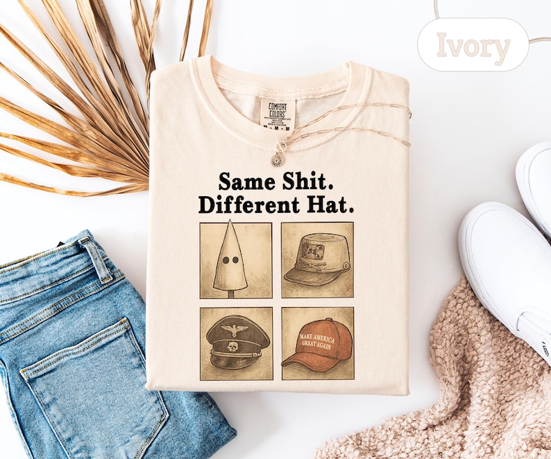 Peut inclure: T-shirt de couleur ivoire avec l'inscription "Same Shit. Different Hat." et quatre illustrations de chapeaux. Le t-shirt est pos&eacute; &agrave; plat avec un jean bleu, des chaussures blanches et une plante d&eacute;corative en arri&egrave;re-plan.