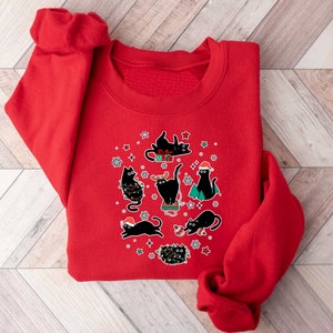 May include: Red Christmas sweatshirt featuring a design of black cats. The cats are wearing Santa hats and are surrounded by Christmas lights and decorations.