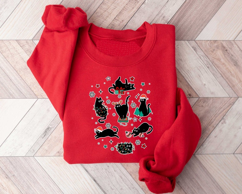 May include: Red Christmas sweatshirt featuring a design of black cats. The cats are wearing Santa hats and are surrounded by Christmas lights and decorations.
