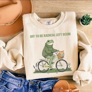 May include: Cream-colored sweatshirt with the text "OFF TO BE RADICAL LEFT SCUM." The graphic depicts a frog riding a bicycle with a cat in a basket. The sweatshirt is paired with blue jeans, a hat, and sandals.