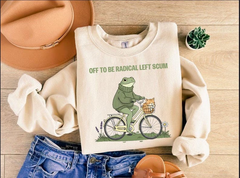 May include: Cream-colored sweatshirt with the text "OFF TO BE RADICAL LEFT SCUM." The graphic depicts a frog riding a bicycle with a cat in a basket. The sweatshirt is paired with blue jeans, a hat, and sandals.