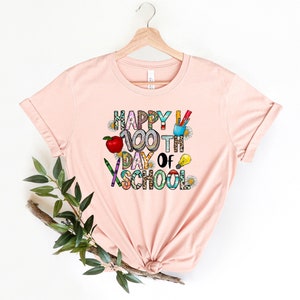 Puede incluir: Una camiseta rosa con el texto "Happy 100th Day of School" en letras de colores y gr&aacute;ficos de tem&aacute;tica escolar.