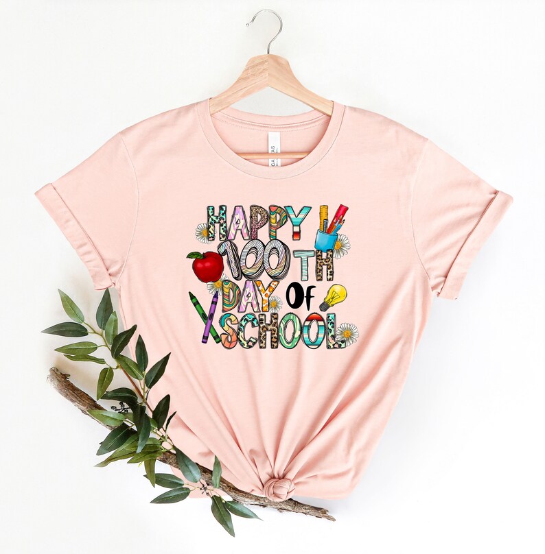Puede incluir: Una camiseta rosa con el texto "Happy 100th Day of School" en letras de colores y gr&aacute;ficos de tem&aacute;tica escolar.