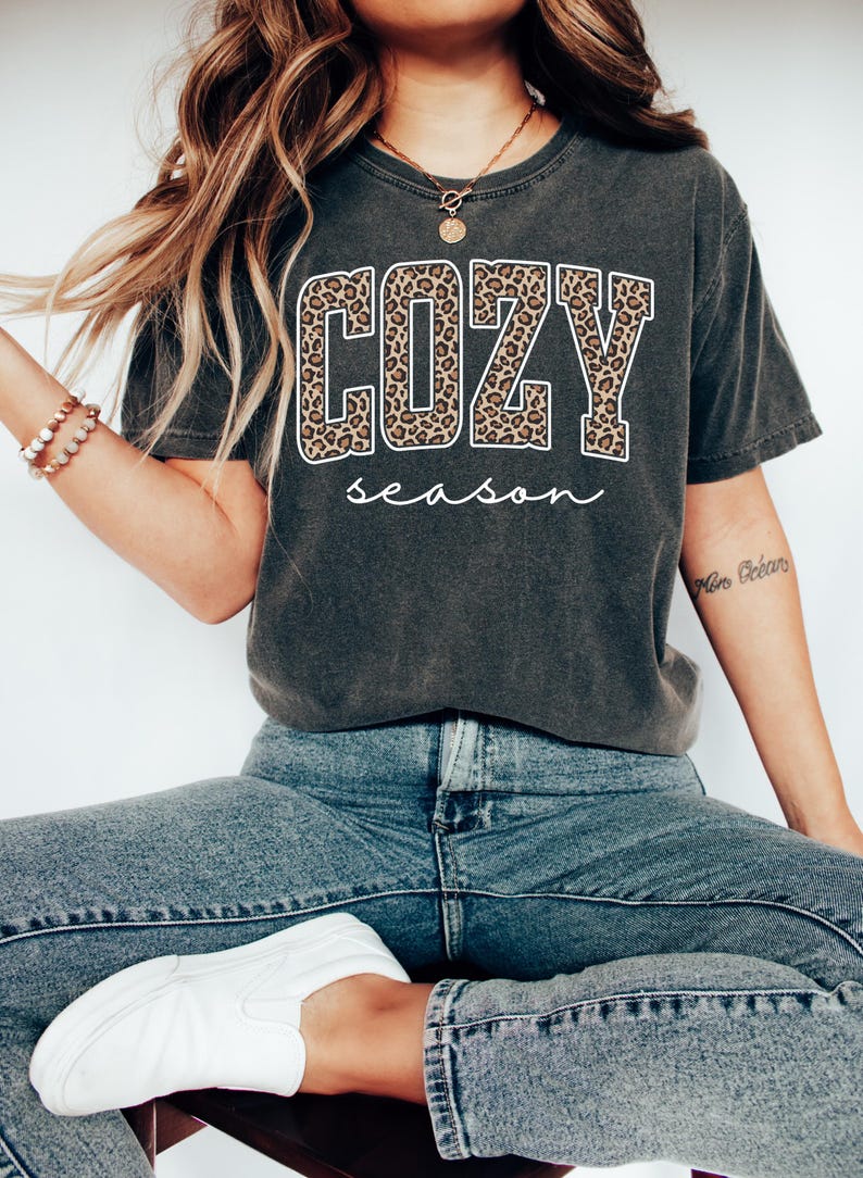 Pu&ograve; includere: T-shirt grigio scuro con le parole "COZY season" stampate sul davanti. La parola "COZY" &egrave; in grandi lettere con un motivo leopardato. La maglietta &egrave; abbinata a jeans blu e scarpe bianche.