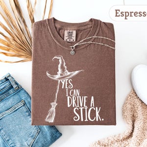 May include: Espresso-colored t-shirt with a witch hat and broom graphic. The text reads "YES I CAN DRIVE A STICK." A silver necklace and white sneakers are also visible. The shirt is paired with blue jeans and a beige scarf.