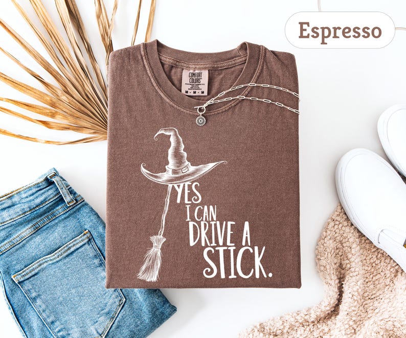 May include: Espresso-colored t-shirt with a witch hat and broom graphic. The text reads "YES I CAN DRIVE A STICK." A silver necklace and white sneakers are also visible. The shirt is paired with blue jeans and a beige scarf.