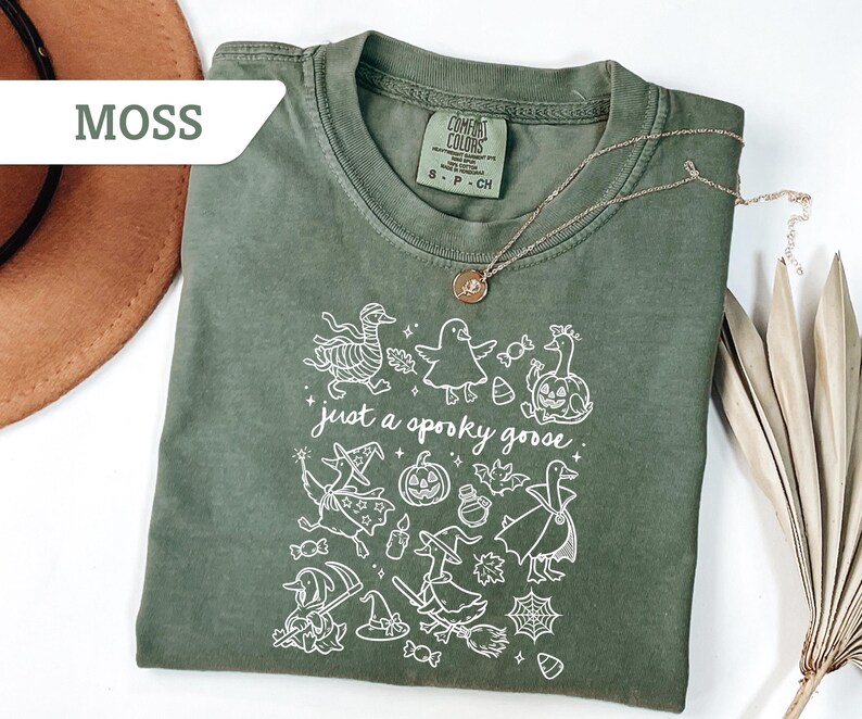 May include: Moss green t-shirt with a white graphic of Halloween-themed geese and the phrase "just a spooky goose." The shirt has a crew neck and short sleeves. A gold necklace with a pendant is draped across the shirt.