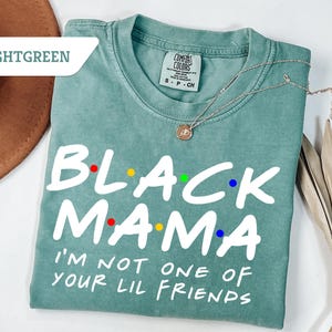 May include: Light green t-shirt featuring the text "BLACK MAMA" in white, with colourful dot accents. The shirt also reads "I'M NOT ONE OF YOUR LIL FRIENDS". A silver necklace with a pendant is on the shirt. The shirt is displayed on a white surface.