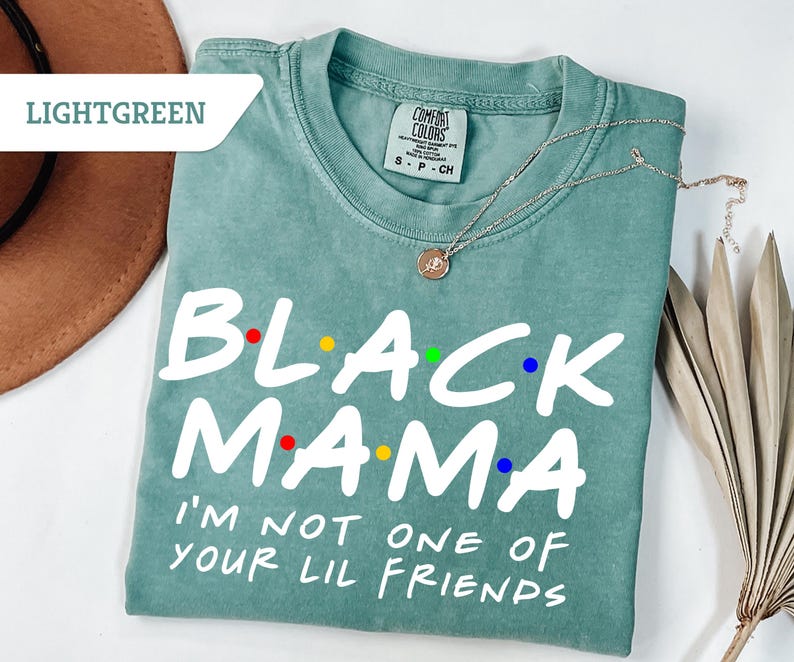 May include: Light green t-shirt featuring the text "BLACK MAMA" in white, with colourful dot accents. The shirt also reads "I'M NOT ONE OF YOUR LIL FRIENDS". A silver necklace with a pendant is on the shirt. The shirt is displayed on a white surface.