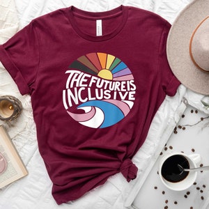 May include: A maroon t-shirt with a circular graphic design featuring a rainbow, sun, and waves. The text "THE FUTURE IS INCLUSIVE" is written in white on the graphic.