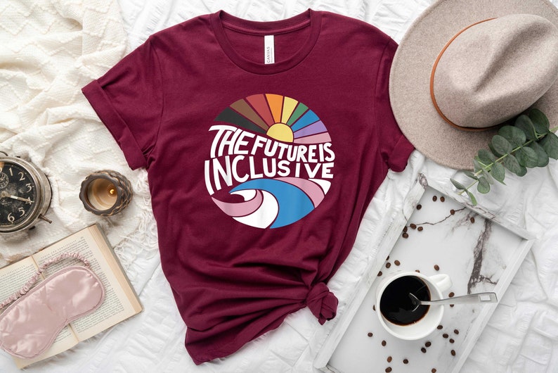 May include: A maroon t-shirt with a circular graphic design featuring a rainbow, sun, and waves. The text "THE FUTURE IS INCLUSIVE" is written in white on the graphic.