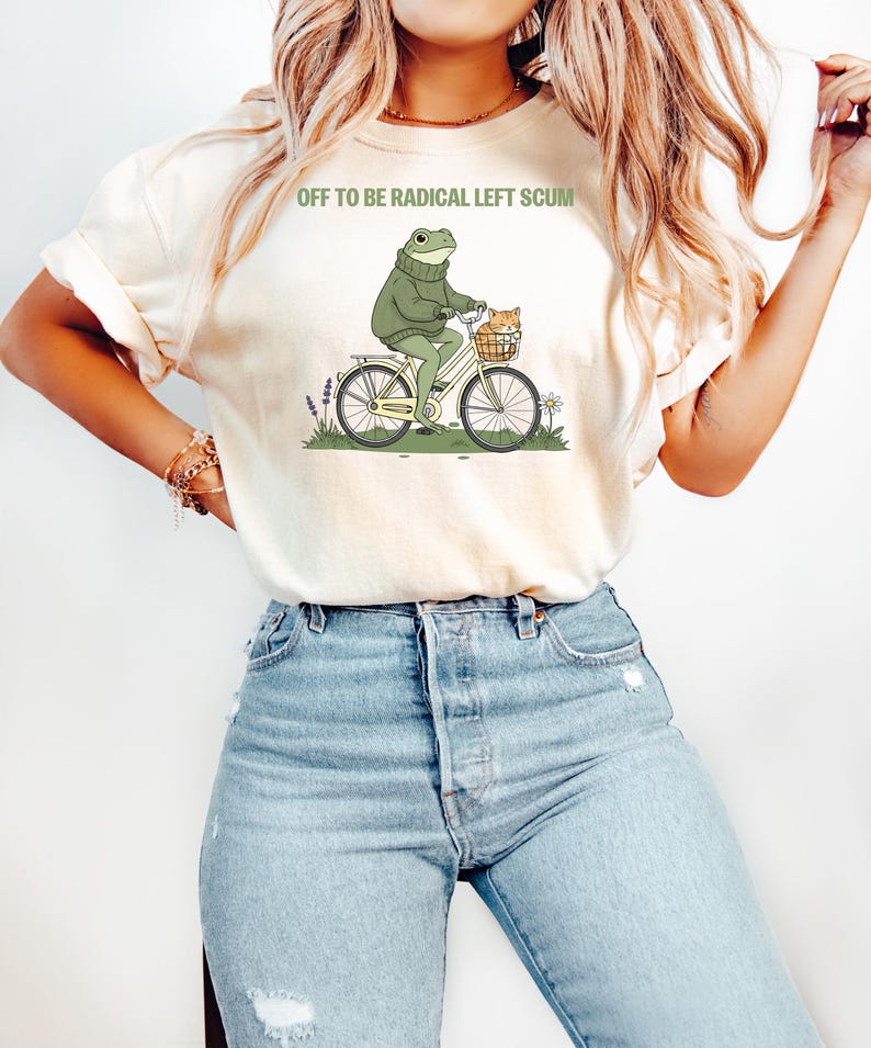 May include: Cream-colored t-shirt with the text "OFF TO BE RADICAL LEFT SCUM." The shirt features a cartoon frog riding a bicycle with a cat in the basket. The frog wears a green sweater.
