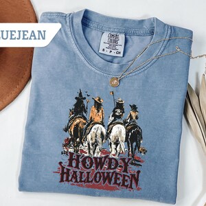 May include: Bluejean Comfort Colors t-shirt featuring a graphic of four figures on horseback, with the text "HOWDY HALLOWEEN" in red. A gold necklace with a pendant is draped on the shirt. A brown hat is visible in the upper left corner.
