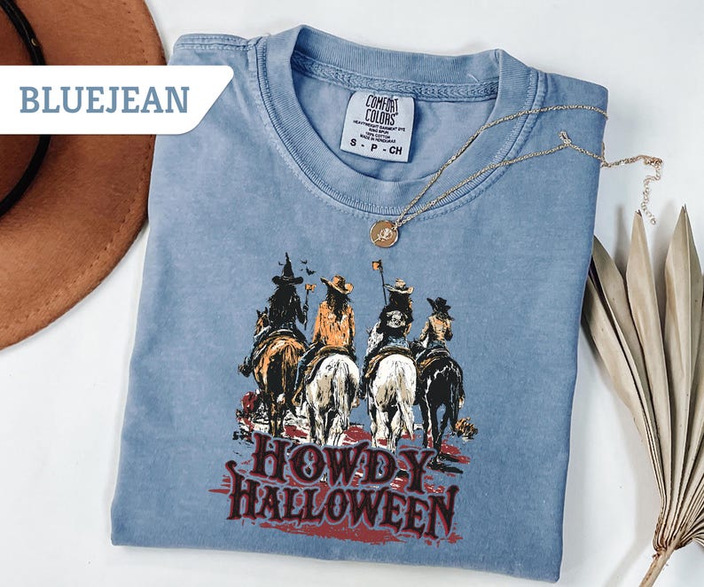 May include: Bluejean Comfort Colors t-shirt featuring a graphic of four figures on horseback, with the text "HOWDY HALLOWEEN" in red. A gold necklace with a pendant is draped on the shirt. A brown hat is visible in the upper left corner.