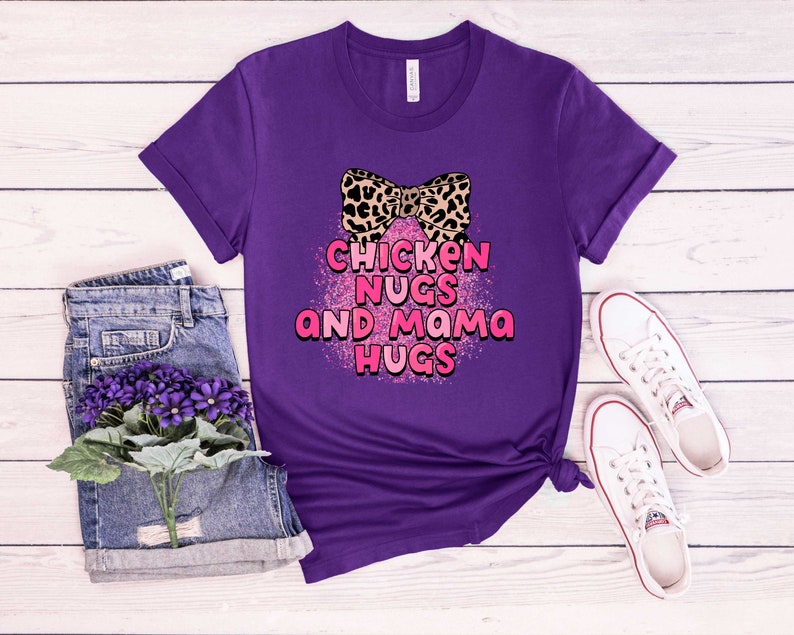 May include: Purple t-shirt featuring a pink and black leopard print bow and the text "Chicken Nugs and Mama Hugs" in pink lettering with a pink glitter effect.