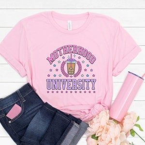 May include: Pink t-shirt with the text "Motherhood University" in a pink and purple font. A cartoon image of a boba tea cup is in the center of the text. The shirt is on a white wooden surface with a pink tumbler and flowers.