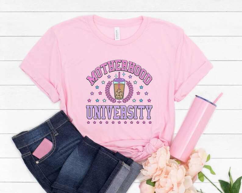 May include: Pink t-shirt with the text "Motherhood University" in a pink and purple font. A cartoon image of a boba tea cup is in the center of the text. The shirt is on a white wooden surface with a pink tumbler and flowers.