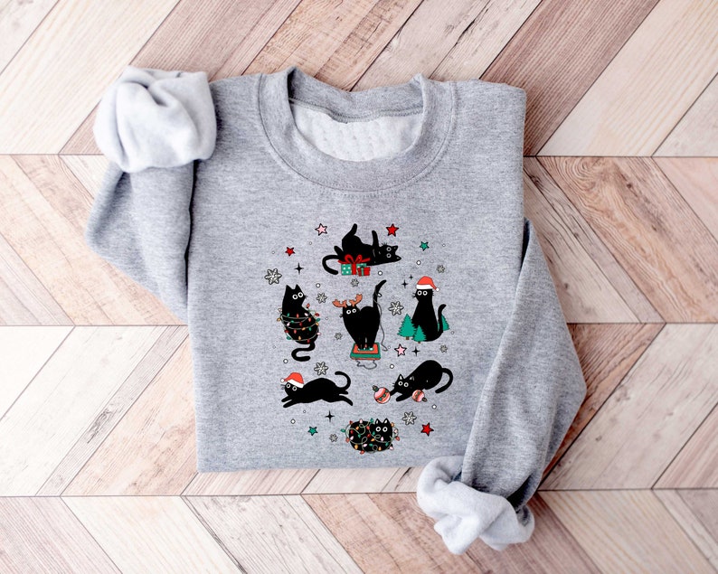 May include: A grey sweatshirt with a Christmas-themed design featuring black cats wearing Santa hats and holding presents. The cats are surrounded by stars, snowflakes, and Christmas lights.