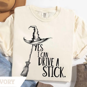 May include: Ivory-colored t-shirt with a black graphic of a witch's hat and broom. The text "YES I CAN DRIVE A STICK" is printed below the hat. The shirt is a Comfort Colors brand.