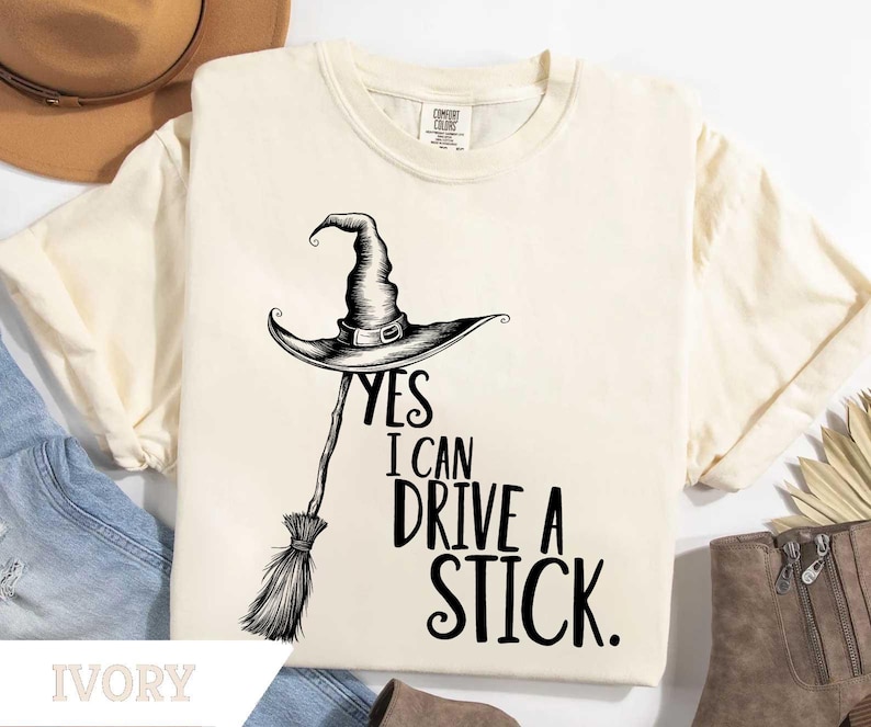 May include: Ivory-colored t-shirt with a black graphic of a witch's hat and broom. The text "YES I CAN DRIVE A STICK" is printed below the hat. The shirt is a Comfort Colors brand.
