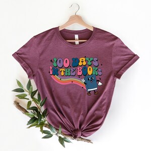 May include: A maroon t-shirt with a graphic design of a book character with the text "100 Days in the Books" in a rainbow font.