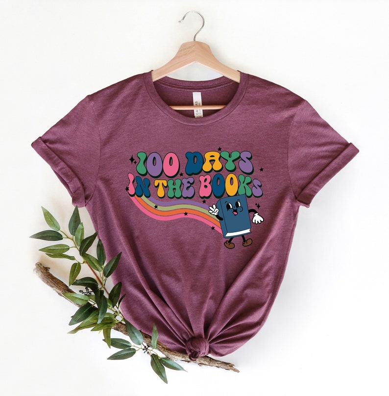May include: A maroon t-shirt with a graphic design of a book character with the text "100 Days in the Books" in a rainbow font.