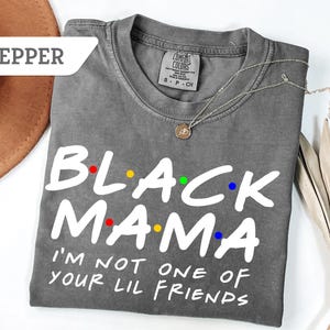 May include: A grey t-shirt with the words "BLACK MAMA" in white, with colourful dots above the letters. Below the text is "I'M NOT ONE OF YOUR LIL FRIENDS" in white. A silver necklace with a pendant is on the shirt.