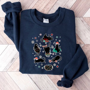 May include: A navy blue sweatshirt with a Christmas-themed design featuring black cats wearing Santa hats and holding presents. The cats are surrounded by snowflakes, stars, and Christmas lights.