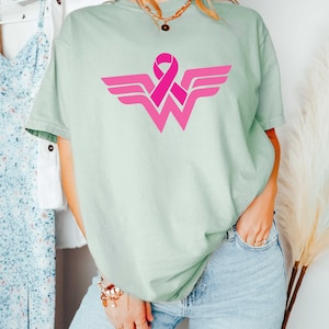 May include: A light green t-shirt with a pink ribbon design in the shape of the Wonder Woman logo.
