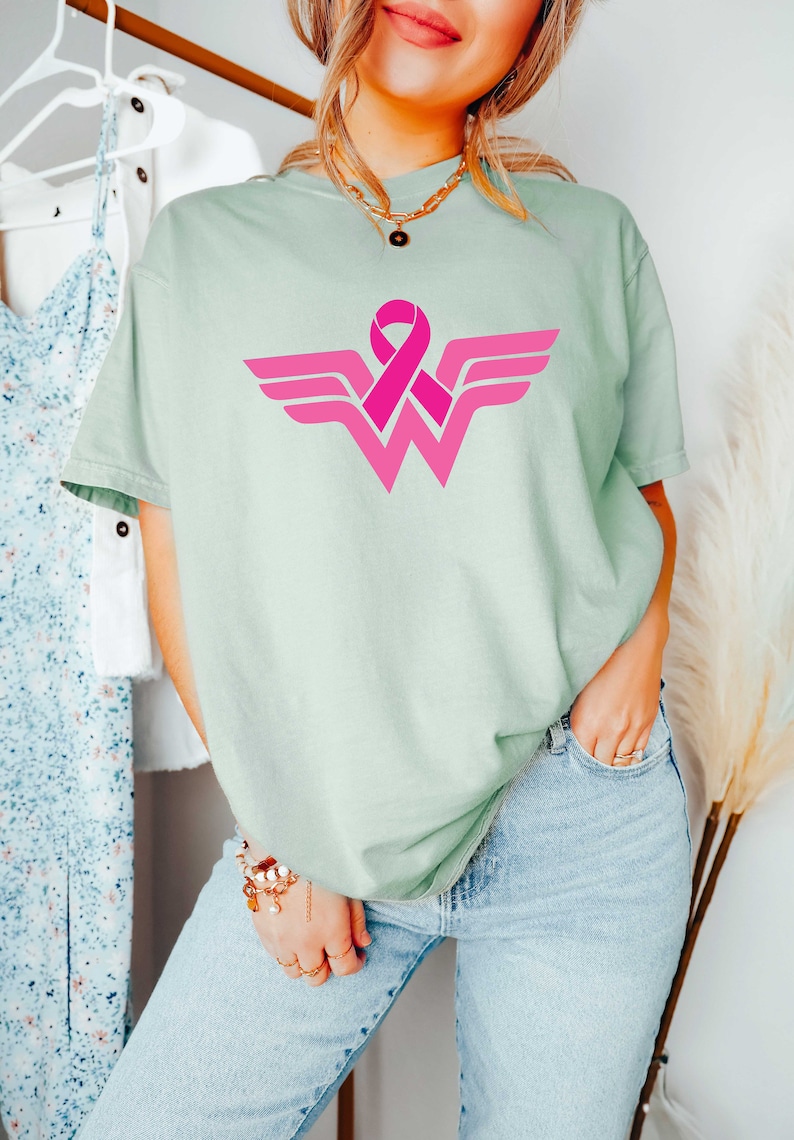 May include: A light green t-shirt with a pink ribbon design in the shape of the Wonder Woman logo.