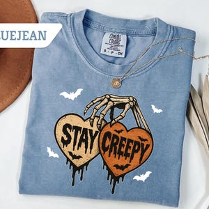 May include: Bluejean Comfort Colors t-shirt with a skeleton hand holding two heart-shaped pumpkins that read "Stay Creepy". The shirt has a crew neck and features white bat graphics. A gold necklace is draped across the shirt.