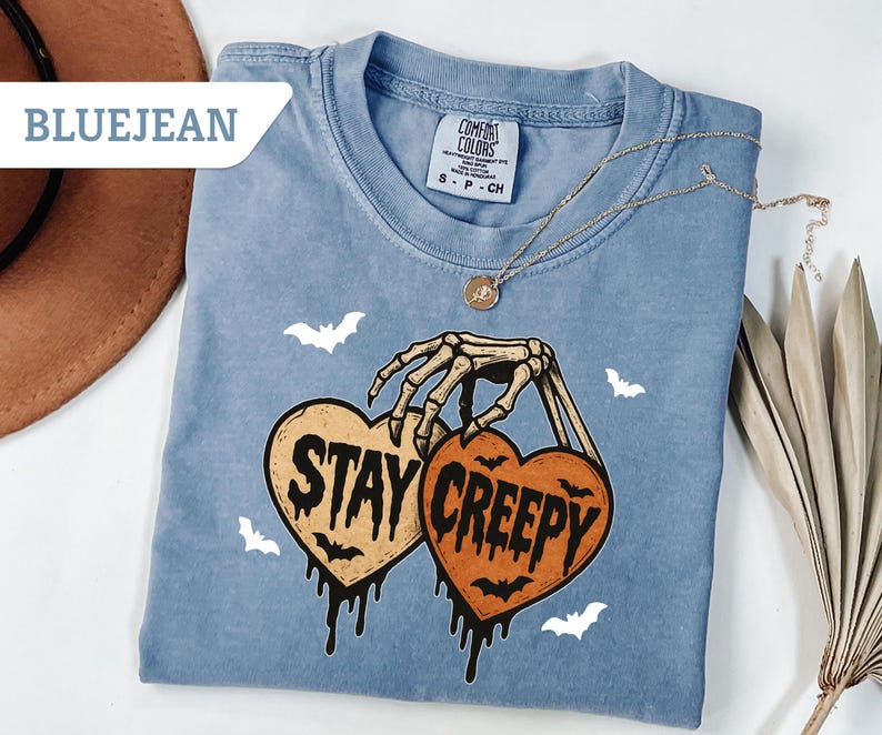May include: Bluejean Comfort Colors t-shirt with a skeleton hand holding two heart-shaped pumpkins that read "Stay Creepy". The shirt has a crew neck and features white bat graphics. A gold necklace is draped across the shirt.