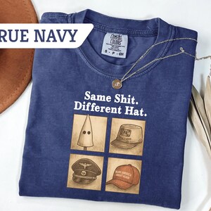 Peut inclure: T-shirt bleu marine avec l'inscription "Same Shit. Different Hat." et quatre illustrations de chapeaux. Un collier dor&eacute; et un chapeau marron sont &eacute;galement visibles.