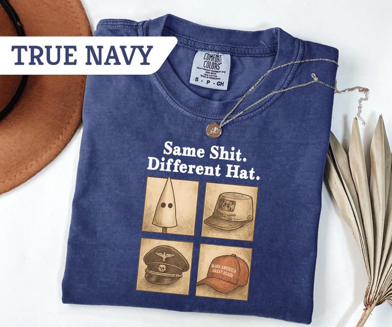 Peut inclure: T-shirt bleu marine avec l'inscription "Same Shit. Different Hat." et quatre illustrations de chapeaux. Un collier dor&eacute; et un chapeau marron sont &eacute;galement visibles.