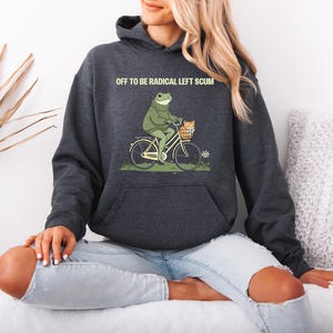May include: Dark gray hoodie with a frog riding a bicycle, carrying a cat in a basket. The text "OFF TO BE RADICAL LEFT SCUM" is printed above the frog. The hoodie has a kangaroo pocket and a drawstring hood.