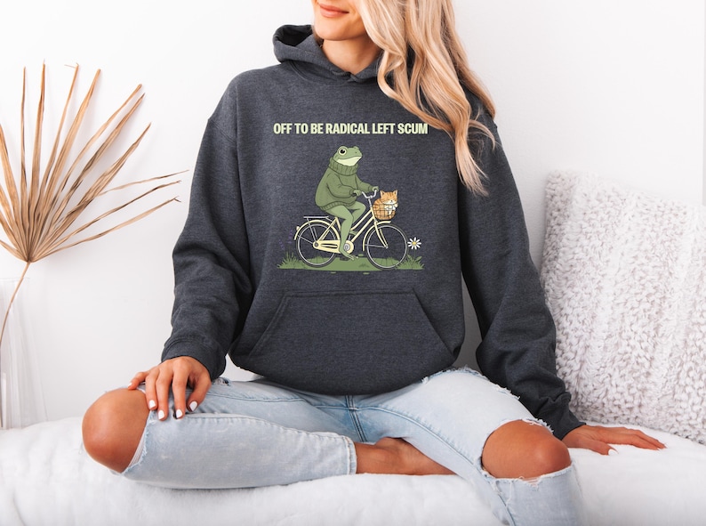 May include: Dark gray hoodie with a frog riding a bicycle, carrying a cat in a basket. The text "OFF TO BE RADICAL LEFT SCUM" is printed above the frog. The hoodie has a kangaroo pocket and a drawstring hood.