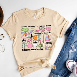 May include: A beige t-shirt with a colorful graphic design featuring the word "OT" surrounded by various icons representing occupational therapy skills, such as play, social, sensory processing, and bilateral coordination.