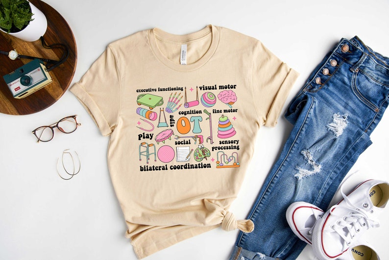May include: A beige t-shirt with a colorful graphic design featuring the word "OT" surrounded by various icons representing occupational therapy skills, such as play, social, sensory processing, and bilateral coordination.
